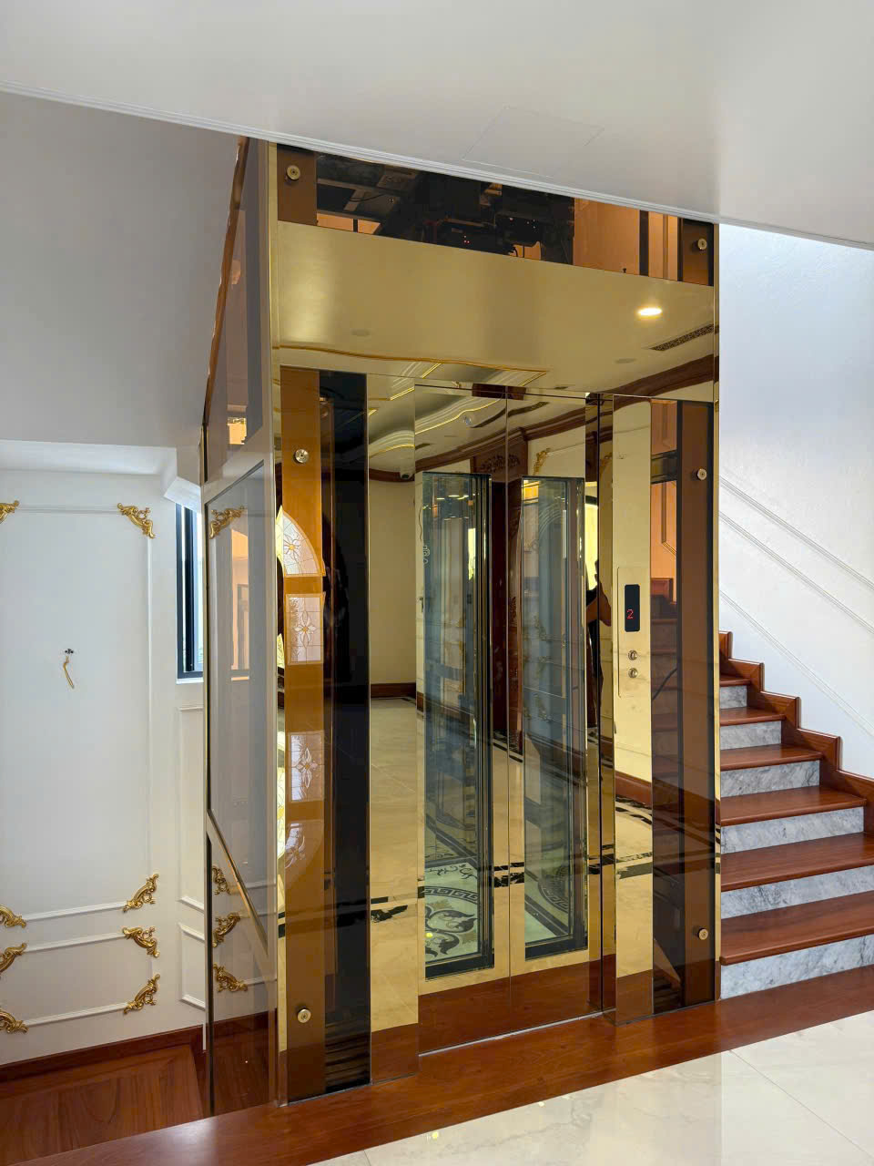 Thang máy kính khung inox mạ vàng lắp trong nhà cạnh cầu thang gỗ marble sang trọng